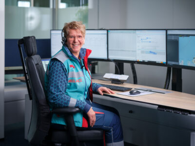 Rita kijkt lachend in de camera terwijl ze achter haar bureau bij de meldkamer zit.