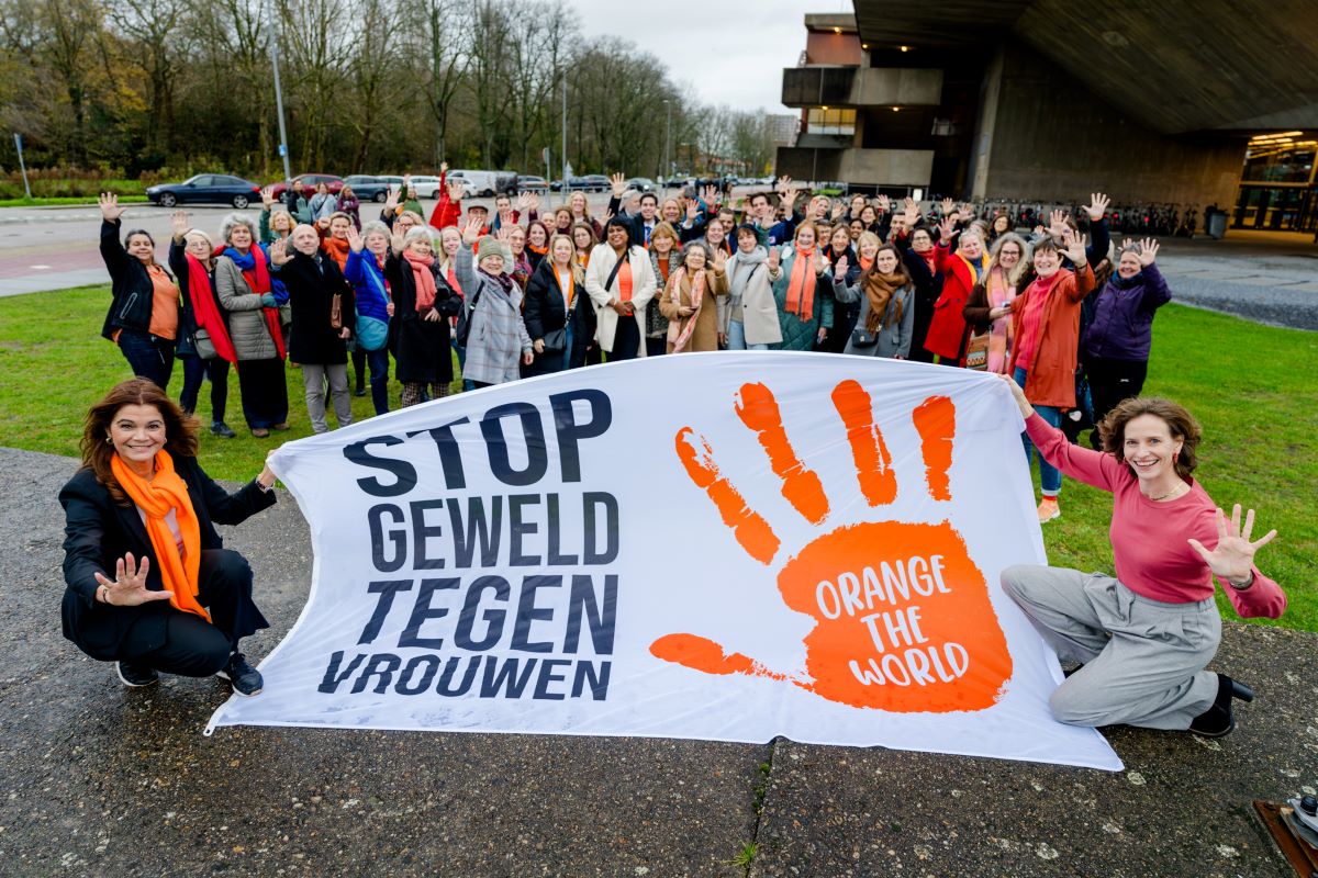 2 vrouwen houden de Orange the World vlag vast met daarop de tekst 'Stop geweld tegen vrouwen'. Daarachter een grote groep van vooral vrouwen.
